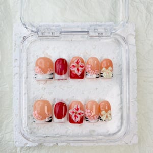 May include: A set of ten press-on nails in a clear plastic case. The designs include solid red nails, nails with pink floral accents, nails with white and red flower art, and nails featuring small cream-colored bows on a nude base with black stripes.