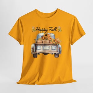May include: A mustard yellow t-shirt featuring a graphic of a vintage pickup truck carrying a wooden crate filled with sunflowers and pumpkins. The text "Happy Fall" is printed in black script above the truck, flanked by small floral accents.