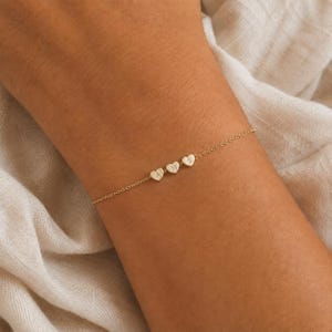 Könnte beinhalten: Ein zartes Goldkettenarmband mit drei kleinen, herzförmigen Anhängern, die in einer Reihe angeordnet sind. Jeder Herzanhänger ist mit einem einzelnen Großbuchstaben graviert, nämlich den Buchstaben A, M und S, die mittig an der Kette sitzen.