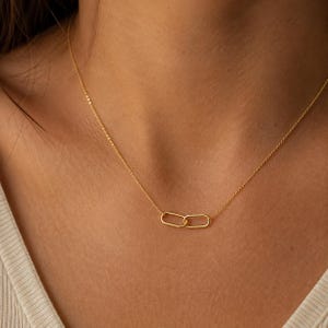 Könnte beinhalten: Eine goldfarbene Halskette mit einer feinen Kette und einem zentralen Anhänger aus zwei ineinandergreifenden ovalen Gliedern. Die Kette liegt auf der Haut und zeigt ein schlichtes, raffiniertes Design mit einer polierten Metalloberfläche.