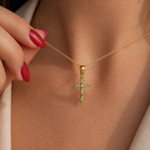 Könnte beinhalten: Eine goldene Halskette mit einem Kreuzanhänger. Das Kreuz besteht aus vier hellgrünen Steinen im Marquise-Schliff, die in einer vertikalen und horizontalen Anordnung gesetzt sind, mit einem kleinen klaren runden Stein in der Mitte.