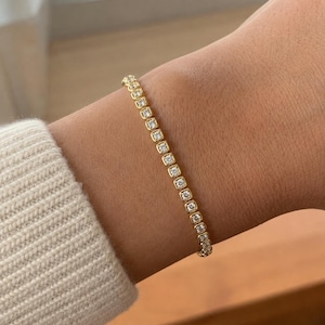 Könnte beinhalten: Ein goldenes Tennisarmband mit einer durchgehenden Reihe quadratischer Glieder, die jeweils mit einem einzelnen runden, klaren Edelstein besetzt sind. Das Armband hat eine polierte metallische Oberfläche und ein flexibles Design.