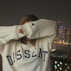May include: An off-white crewneck sweatshirt featuring the word DISSENT in bold black collegiate-style lettering across the chest. The sleeves have a small black star embroidered on the cuff. The fabric appears to be a soft, thick cotton blend.