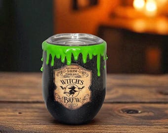 May include: A black stemless wine tumbler with a green, dripping rim. The tumbler features a label that reads "Witch's Brew" with a witch silhouette. The background is a blurred wooden surface.