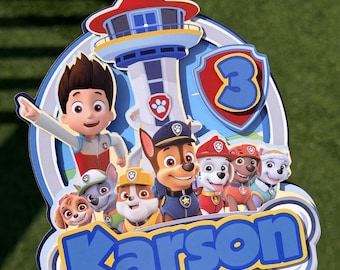 May include: A multi-layered cake topper featuring the Paw Patrol characters and the Lookout Tower. The design includes the name Karson in blue letters, the number 3 in a red shield, and a clear plastic stick at the bottom for cake insertion.