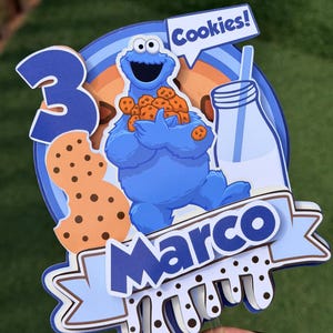 Könnte beinhalten: Ein mehrschichtiger Tortenaufleger aus Papier mit einem blauen Monster, das einen Haufen Kekse hält, einem Glas Milch, einer großen Zahl 3 und einem Banner mit dem Namen Marco. Das Design enthält eine Sprechblase mit dem Text Cookies! in Blau und Weiß.