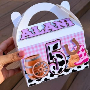 Puede incluir: Una caja de cartón blanca con asa que muestra el nombre ALANI en letras moradas. La caja está decorada con un patrón de cuadros vichy rosa, un barril de madera marrón, una rueda de carro, el número 5 con estampado de vaca, una herradura y una bota.