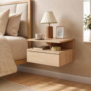 Puede incluir: Una mesita de noche flotante de madera clara con una superficie plana, un estante de almacenamiento abierto y un cajón. La mesita está montada en la pared y sostiene una lámpara de mesa con pantalla plisada, una taza de cerámica, un cuadro y un reloj.
