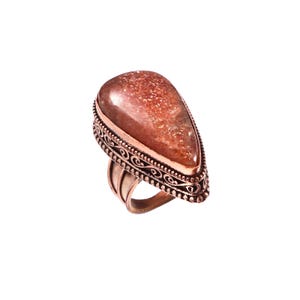 Peut inclure: Une bague en pierre de soleil en forme de goutte sertie sur une bande en cuivre. La pierre précieuse présente une teinte brun rougeâtre avec des inclusions scintillantes internes. Le sertissage métallique affiche un motif de volutes en relief.