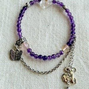 May include: A beaded bracelet featuring purple amethyst spheres, a clear flower-shaped bead with a red center, and two larger translucent beads. It includes silver-toned metal charms of a butterfly, a stylized animal, and a bell-shaped pendant.