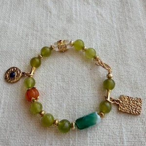 May include: A beaded bracelet featuring round olive green beads, a single orange fluted bead, a cylindrical green and white bead, and a clear bead with gold filigree. The bracelet includes gold-toned metal spacers and two charms: one round with a dark blue center and one rectangular with a textured surface.