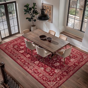 May include: A large rectangular area rug featuring a deep red background with intricate floral and vine patterns in cream and muted tones, positioned under a wooden dining table with six upholstered chairs in a bright dining room.
