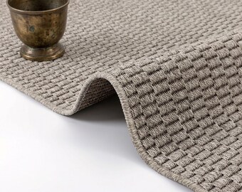 May include: A taupe area rug with a dense, woven basket-weave texture and a finished edge, accompanied by a small, weathered bronze-toned metal chalice or cup resting on the surface of the rug.