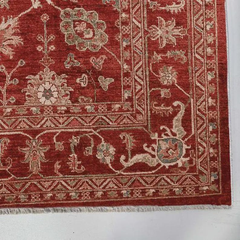 May include: A rectangular area rug featuring a deep red background with intricate floral and vine patterns in muted beige, sage green, and soft blue tones. The rug has a wide decorative border with geometric motifs and a short white fringe.