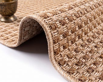 May include: A woven area rug featuring a textured, checkered pattern in shades of tan and light brown. The rug has a finished, bound edge and is folded over to reveal the underside. A small, metallic, pedestal-style cup sits on the rug surface.