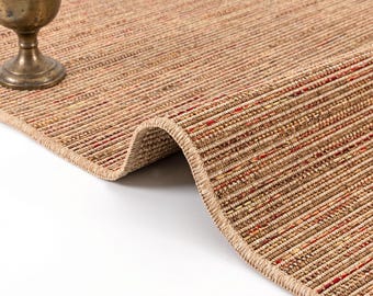 May include: A woven area rug featuring a textured surface with vertical stripes in shades of tan, beige, and brown, accented by intermittent thin lines of red and orange thread. The rug has a finished bound edge and a folded corner.