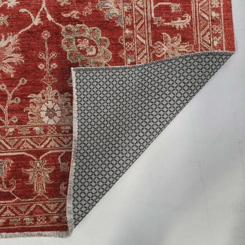 May include: A rectangular area rug featuring a deep red background with intricate beige and light gray floral patterns and geometric borders. The corner is folded back to reveal a black and white non-slip backing with a diamond grid texture.
