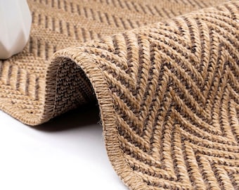 May include: A woven runner rug featuring a tan and brown herringbone pattern. The rug is folded over to reveal the textured, braided construction and the finished stitched edge. The material appears to be a natural fiber, showing a consistent zigzag design throughout the surface of the textile.