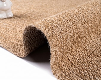 May include: A tan, woven area rug with a thick, textured surface made of natural fibers. The rug is folded over to reveal its dense, braided construction and sturdy backing. A small, white, smooth figurine sits on the corner of the rug.