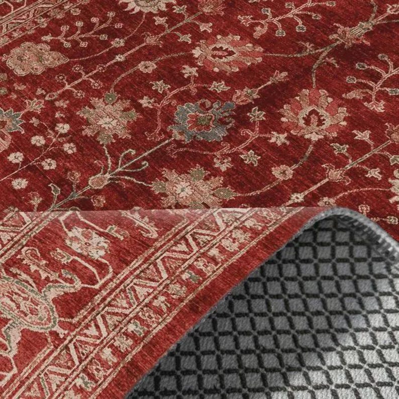May include: A deep red area rug featuring a repeating floral pattern in muted beige, sage green, and slate blue tones. The rug is partially folded over to reveal a dark gray, textured, non-slip backing with a grid-like pattern.