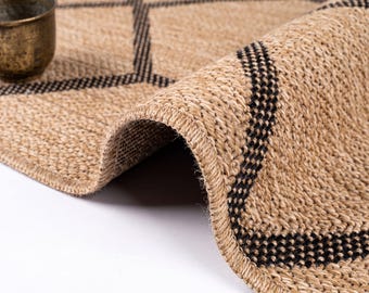 May include: A woven tan jute rug featuring a geometric diamond pattern outlined in black thread. The rug is folded over to show its thick, textured construction, and a small, weathered metallic cup sits on the flat surface of the rug.