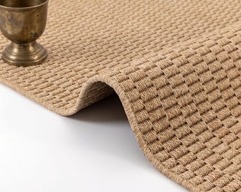 May include: A tan woven area rug with a dense, repeating textured pattern and a finished edge, accompanied by a small, weathered brass chalice-shaped vessel placed on the surface of the rug.
