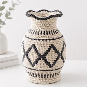 Può includere: Un vaso all'uncinetto con base color crema e un motivo geometrico a rombi grigio scuro al centro. Il vaso presenta un bordo superiore arricciato e strisce orizzontali grigio scuro sul collo e sulla base, realizzato con una trama fitta.