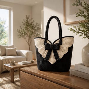 Può includere: Una borsa tote all'uncinetto nera con una grande patta color crema dai bordi smerlati che ricorda delle ali. Un prominente fiocco all'uncinetto nero è centrato sulla patta e la borsa include due robusti manici all'uncinetto neri.