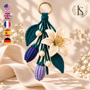 May include: A macrame wall hanging featuring a gold metal ring at the top, from which hang several crocheted elements including a large cream-colored lily, two dark blue buds, one light purple bud, three small white berries, and two dark green leaves.