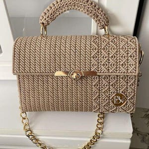 May include: A beige macramé handbag featuring a structured rectangular body with a diagonal cord pattern and a side panel of intricate knotting. It includes a gold-tone metal twist lock closure, a matching top handle, and a gold chain shoulder strap.