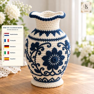 May include: A crochet vase featuring a cream-colored base with intricate dark blue floral and vine patterns. The vase has a ruffled rim and a wide, rounded body that tapers toward the neck and base. The design includes a text overlay indicating a PDF pattern is available in five languages: Nederlands, Français, Deutsch, Italiano, and Español.