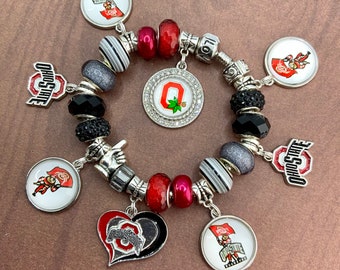 May include: A charm bracelet featuring Ohio State University logos, including the block O with a buckeye leaf, Brutus Buckeye mascot charms, and heart-shaped pendants. The bracelet includes red, black, white, and silver beads with metallic spacers.