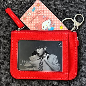 May include: A red rectangular card holder with a clear plastic window displaying a black and white photo card of a person in a shirt and tie. A Hello Kitty card with a pink checkered pattern is tucked inside. It measures 53 by 83 mm.