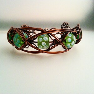 May include: A copper wire bracelet with green and white glass beads. The beads have a polka dot pattern.
