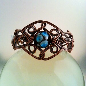 May include: A copper wire bracelet with a blue and white glass bead in the center. The bracelet is made of intricate wirework and has a delicate, feminine design.