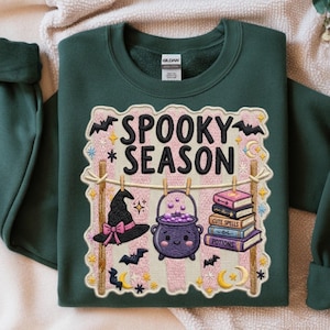 Op de afbeelding: Een donkergroene sweater met ronde hals en een grafische print met de tekst SPOOKY SEASON. Het ontwerp bevat een heksenhoed, een paarse ketel met bubbels en een stapel boeken met de labels CUTE SPELLS en POTIONS aan een waslijn.