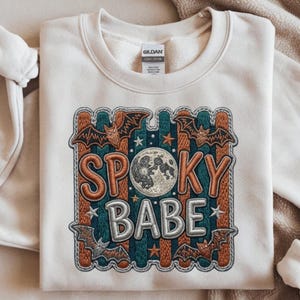 Può includere: Una felpa girocollo color crema con un ricamo che riporta la scritta "SPOOKY BABE". Il disegno include una luna piena, pipistrelli e stelle su uno sfondo a righe verticali arancioni e verde acqua, il tutto racchiuso in un bordo smerlato.