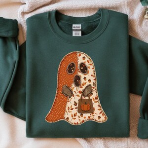 May include: A dark green crewneck sweatshirt featuring a large ghost-shaped appliqué on the chest. The ghost is constructed from two fabric patterns: one side is orange with white polka dots, and the other side features a fall leaf and acorn print.