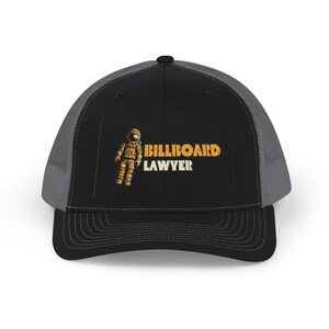 May include: A black and grey trucker hat featuring a graphic of an astronaut in a yellow-orange space suit. To the right of the astronaut, the text BILLBOARD is printed in yellow-orange, with the word LAWYER printed below it in white.