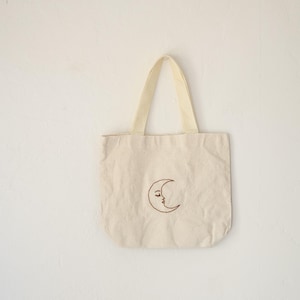 May include: A cream-colored canvas tote bag featuring a brown embroidered crescent moon with a human face. The bag has two matching cream-colored fabric handles attached to the top edge, providing a simple and functional design.