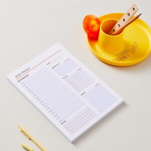 Könnte beinhalten: Ein Tagesplaner-Block mit Zeitplan, Aufgabenliste und Zielen, begleitet von einem gelben Schreibtisch-Organizer mit einem Holzlineal, einem Bleistift, silbernen Büroklammern, einer orangefarbenen Papierdekoration und einem gelben Stift.