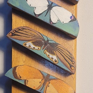May include: A wooden wall hanging featuring eight hand-painted butterfly designs in various colors. Each butterfly is mounted on a curved wooden piece, with colors including yellow, purple, white, orange, pink, blue, and red. The butterflies are arranged vertically on a rectangular wooden board.