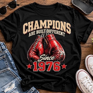 May include: A black t-shirt featuring a graphic of two red boxing gloves in the center. Above the gloves, the text reads "CHAMPIONS ARE BUILT DIFFERENT" in a beige font. Below the gloves, the text reads "Since 1976" with red stars.
