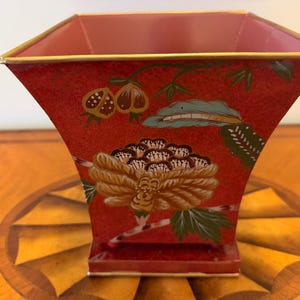 Peut inclure: Un vase en métal de forme carrée avec un bord supérieur évasé et une base étroite. L'extérieur est peint en rouge vif et présente un motif floral détaillé avec des accents dorés, verts et bruns. L'intérieur du vase est d'un rose clair uni.