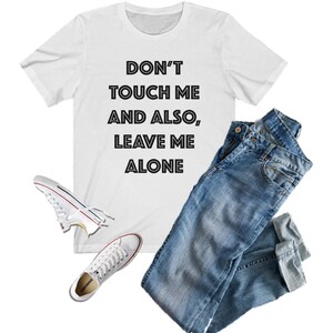 May include: White t-shirt with black text that reads "Don't touch me and also, leave me alone". The shirt is paired with blue jeans and white sneakers.