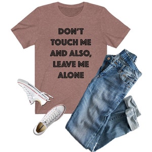 May include: A heather brown t-shirt with the text "Don't touch me and also, leave me alone" in black. The shirt is paired with blue jeans and white sneakers.