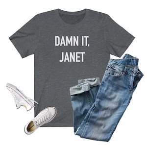 May include: A heather gray t-shirt with the text "DAMN IT, JANET" printed in white. The shirt is paired with blue jeans and white sneakers.