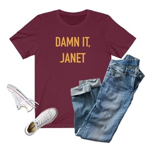 May include: A maroon t-shirt with the text "DAMN IT, JANET" in yellow lettering. The t-shirt is paired with blue jeans and white sneakers.