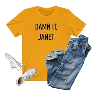 May include: A yellow t-shirt with the text "DAMN IT, JANET" printed in black. The shirt is paired with blue jeans and white sneakers.