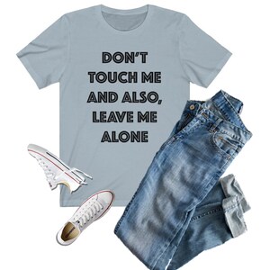 May include: A light blue t-shirt with the text "DON'T TOUCH ME AND ALSO, LEAVE ME ALONE" in black. The shirt is paired with blue jeans and white sneakers.